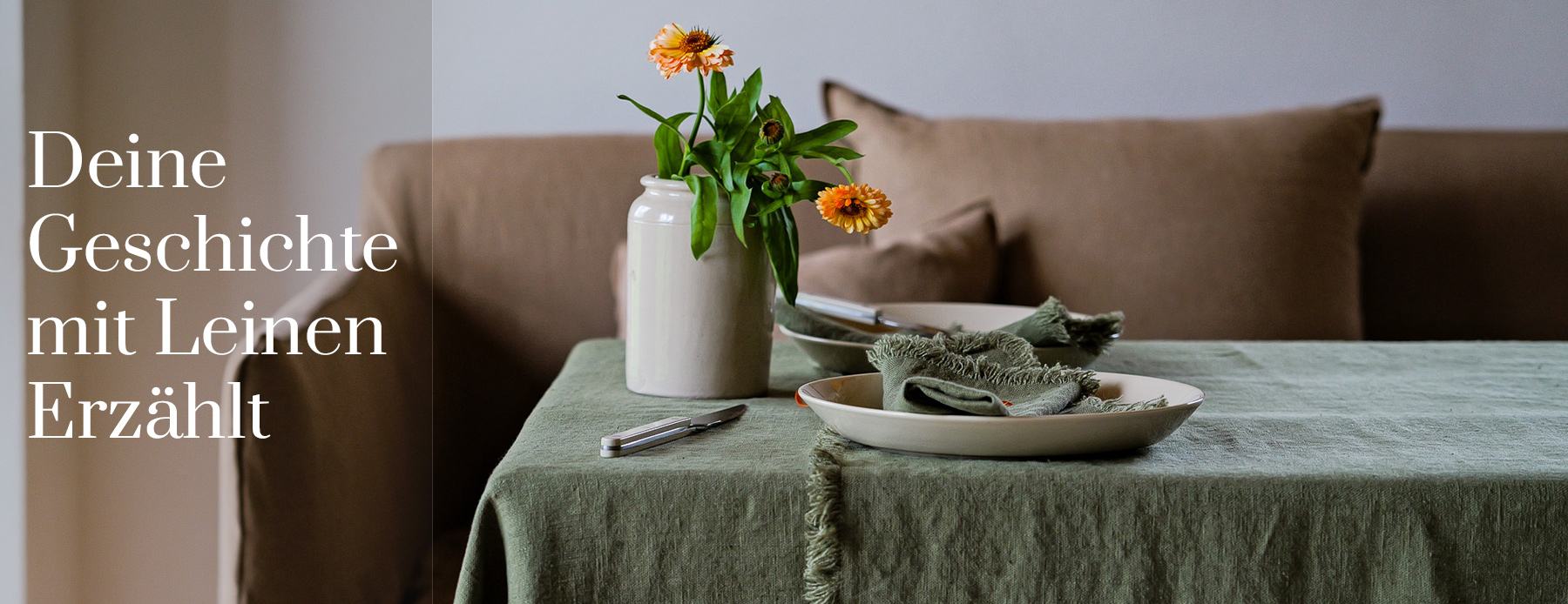 Deine Geschichte mit Leinen erzählt – grüne Leinentischdecke mit Servietten und Herbstblumen vor einem Sofa.