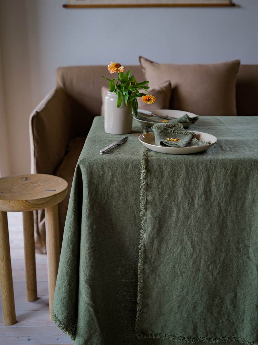 Olivgrüne Leinentischdecke mit Fransen, cremefarbenen Tellern und Blumenvase