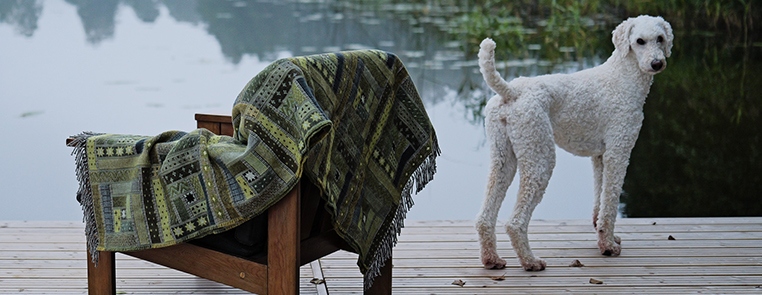 Patterned wool throws draped over a wooden chair beside a misty lake, with a white dog on the jetty.